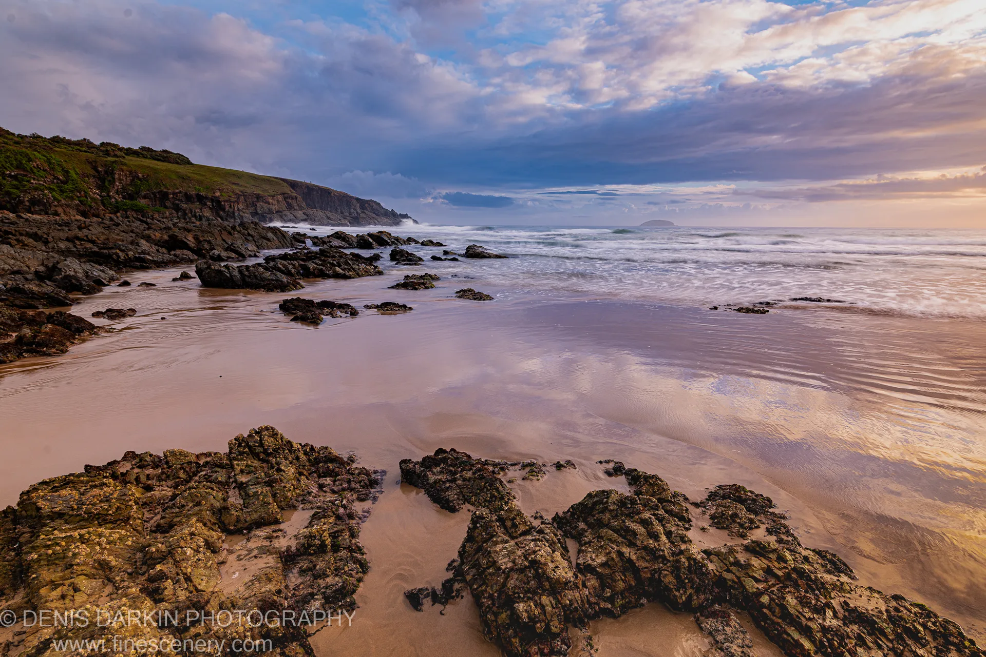 A grassy cliff slopes down to the shore where foam rolls onto moss-covered rocks and wet sand reflects the dawn sky.