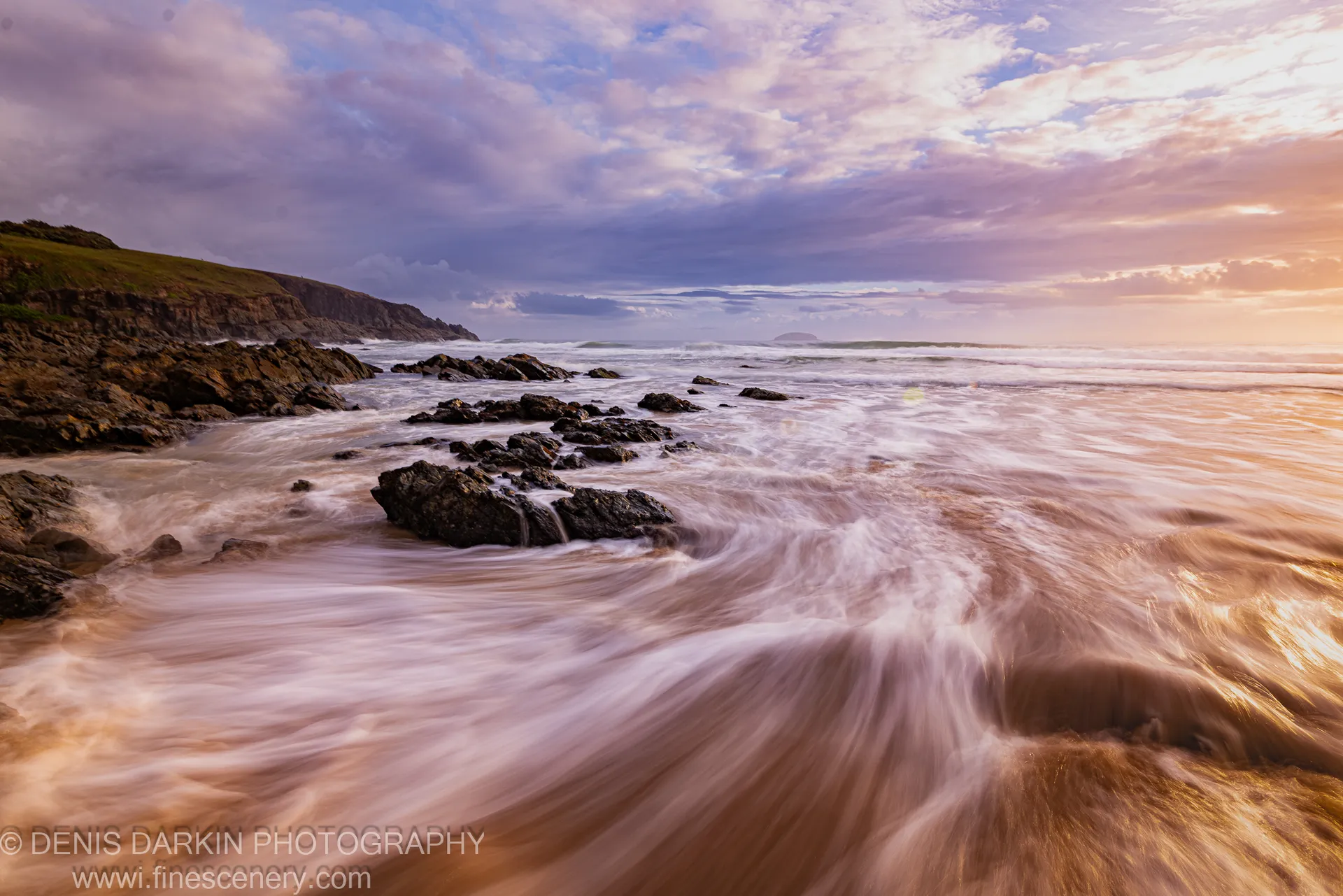 Long-exposure water flows past jagged volcanic rocks on the Coffs Harbor coast at dawn, with a shadowed cliff face anchoring the left side.