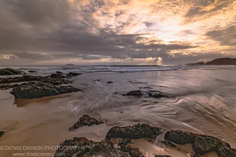 Dark moss-covered rock formations at dawn on the Coffs Harbor coast, with crepuscular rays breaking through heavy storm clouds over the ocean.