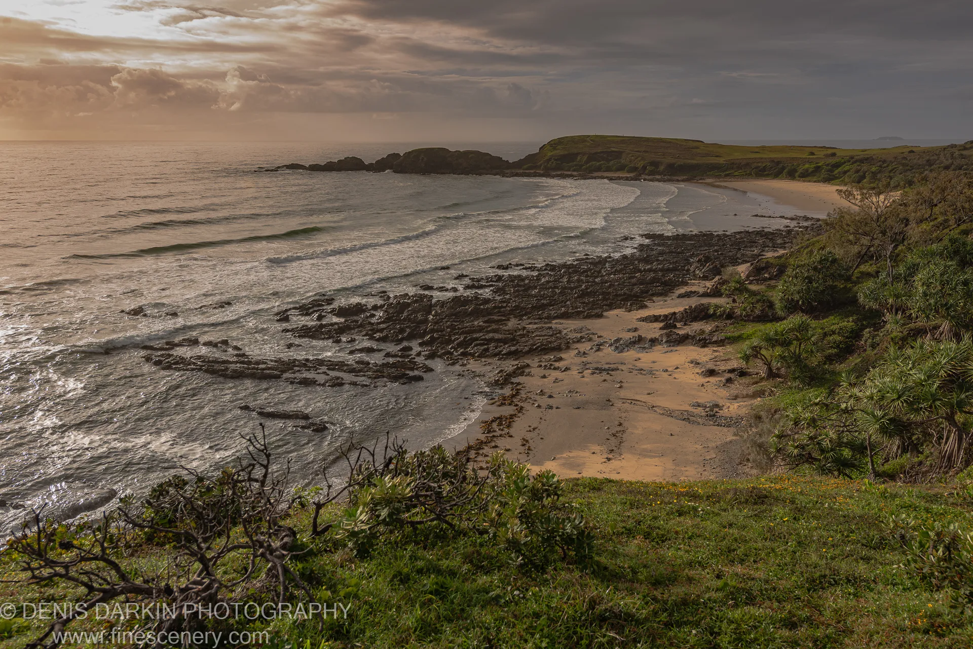 Gentle waves roll onto a curved sandy beach at Coffs Harbor, bordered by dark rocky outcrops and coastal scrub under an overcast morning sky.