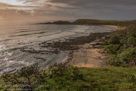 Gentle waves roll onto a curved sandy beach at Coffs Harbor, bordered by dark rocky outcrops and coastal scrub under an overcast morning sky.