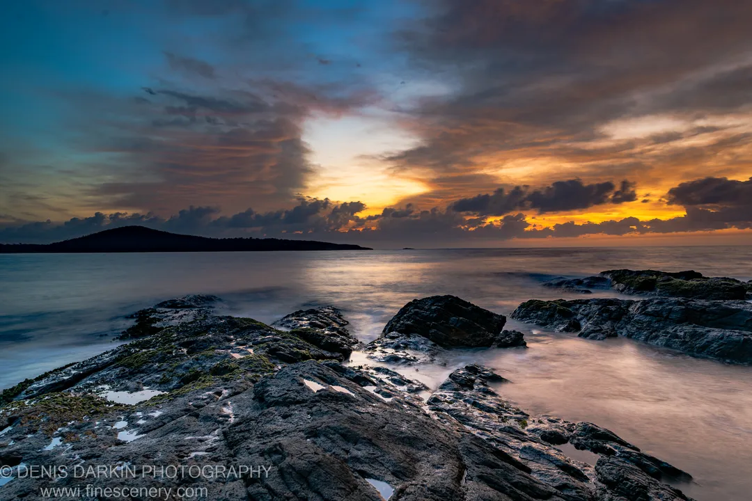 Moss-covered rocks and tide pools along the Nelson Bay coastline at dawn, long-exposure water softened against a warm-toned sunrise sky.