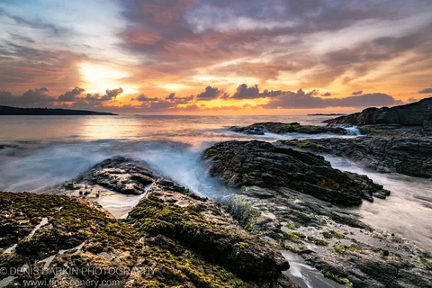 Dark basalt rock formations on the NSW coast at dawn, with long-exposure waves swirling below a cloud-streaked sunrise sky.