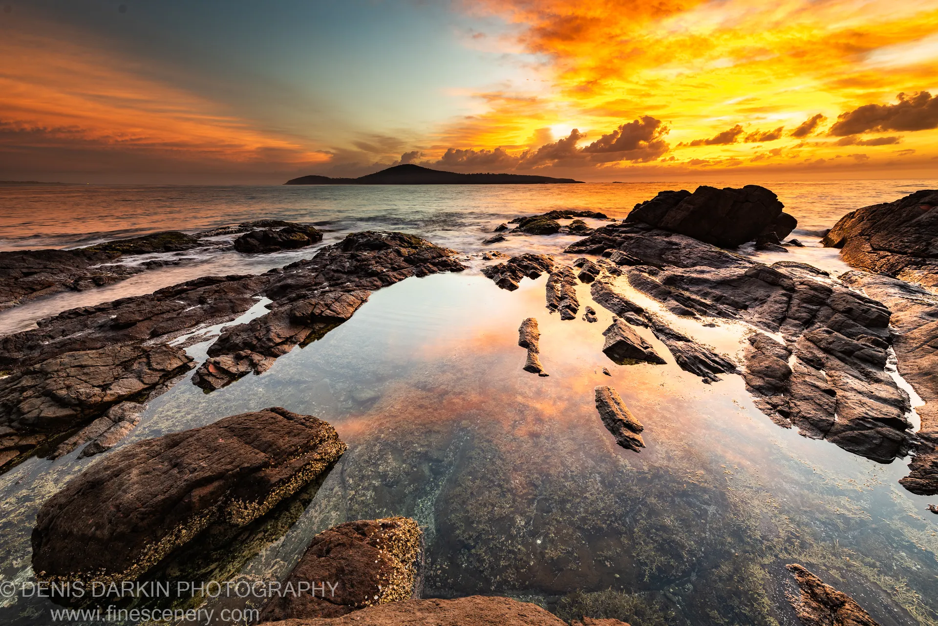Dark rock formations surrounding a tidal pool at dawn on the New South Wales coast, with an island silhouetted against the sunrise sky.
