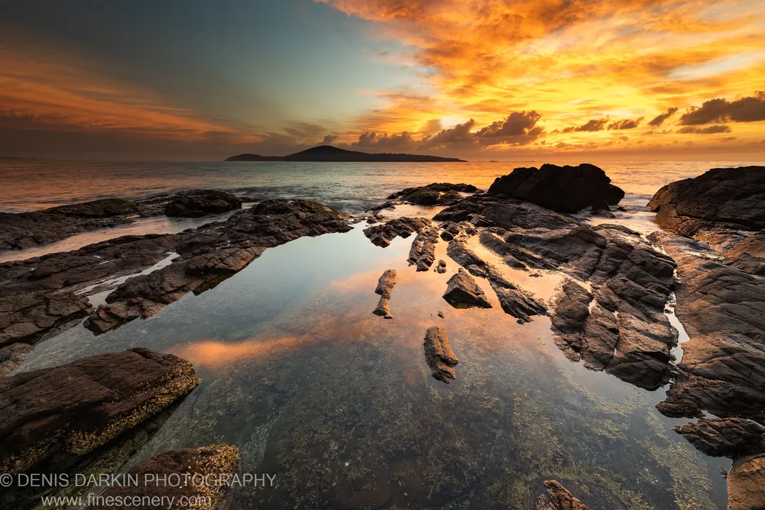 A still tidal pool on basalt rock at dawn, reflecting the orange and blue sunrise sky with an island silhouetted in the distance.