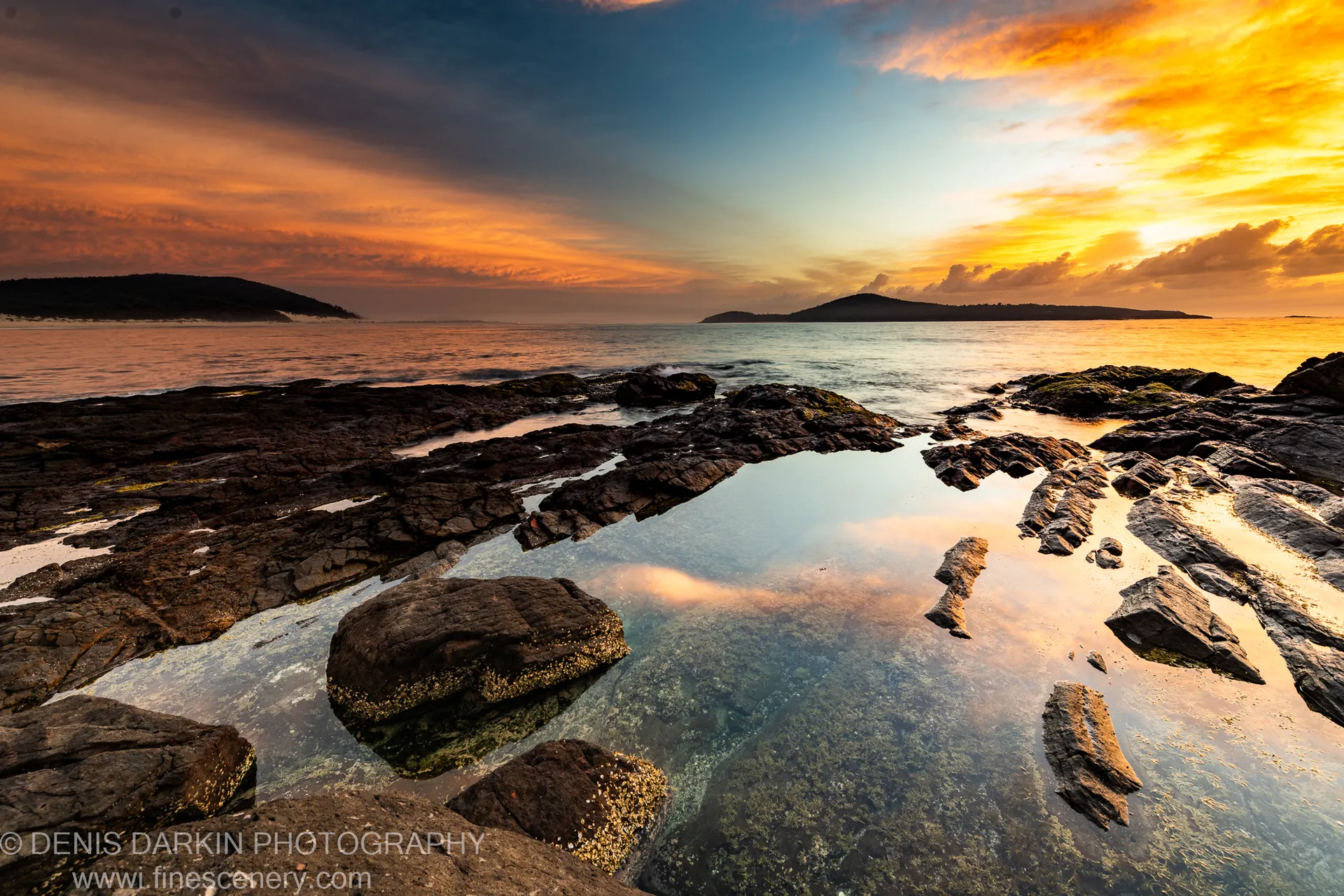 A small tidal pool among dark rocks at dawn near Nelson Bay, reflecting warm orange and yellow sunrise light with distant hills on the horizon.