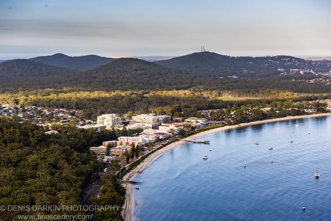 Dozens of boats anchor in the calm cove at Nelson Bay under soft morning light, with buildings along the beach and forested hills behind.
