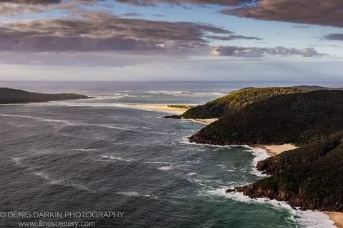 Wide coastal bay view at Nelson Bay with steep forested headlands on either side, waves breaking against dark rocks, and sunlit ridges above shadowed cliffs.