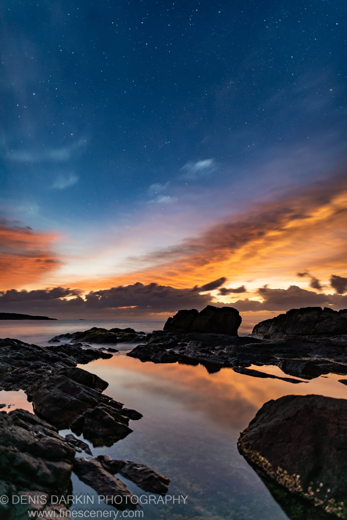 A starry pre-dawn sky over orange horizon light, dark textured rocks and a still pool on the New South Wales coast.