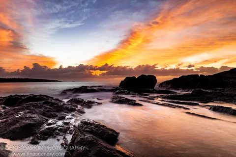 Silhouetted rock formations on the New South Wales coast at dawn, surrounded by long-exposure water while cloud-streaked sky transitions from deep blue to orange above.