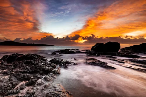 Jagged coastal rock formations at dawn near Nelson Bay, long-exposure water at their base and clouds lit in orange and violet tones above.