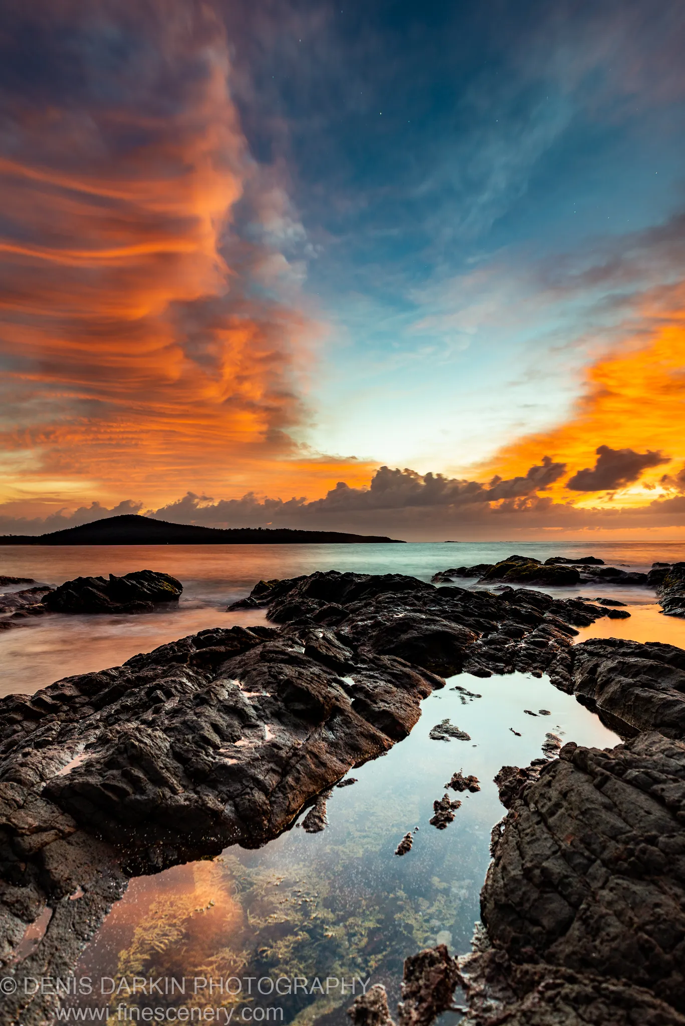 Dark volcanic rocks and a still tidal pool reflect the orange and blue clouds of dawn along the Nelson Bay coast.