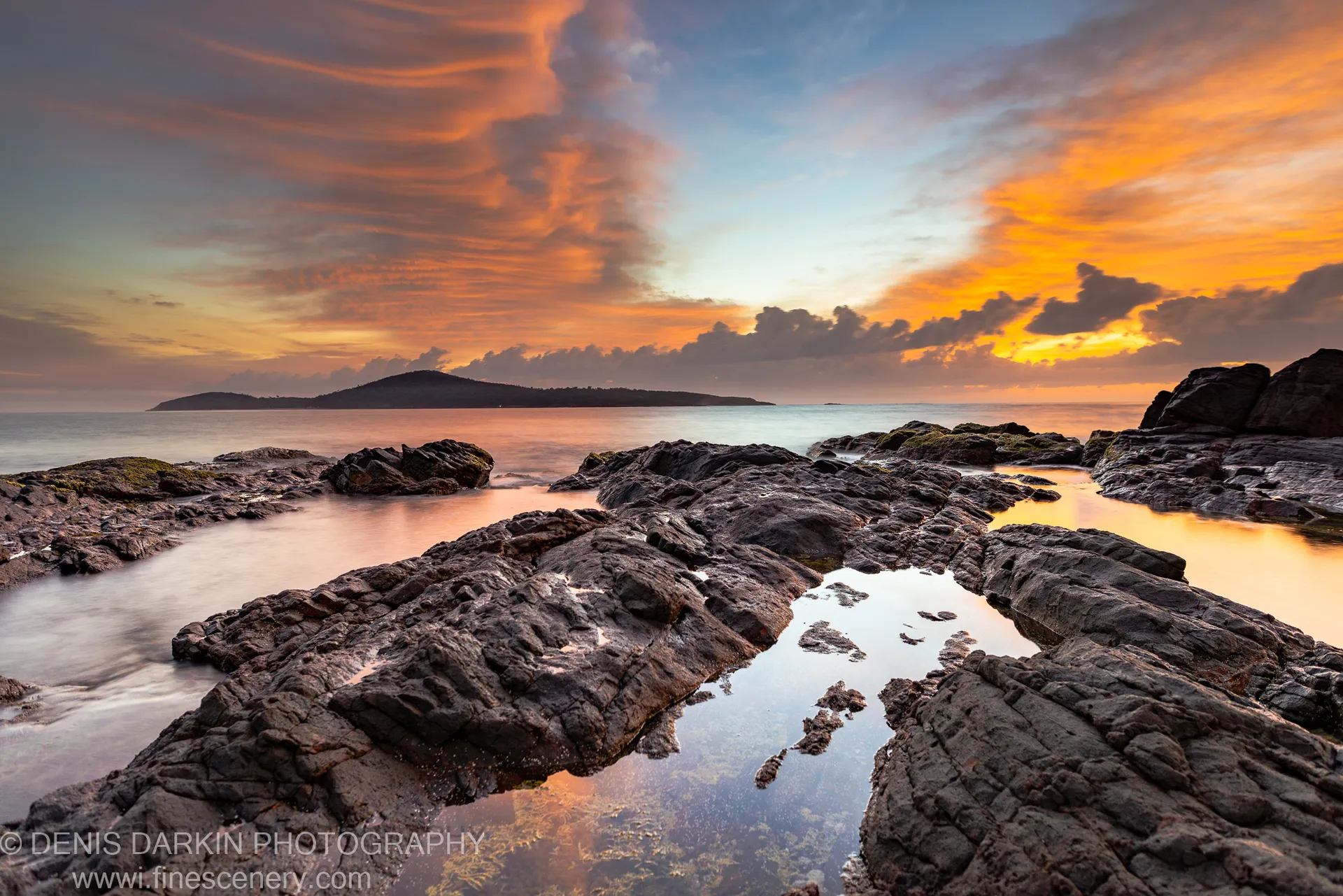 Long-exposure waves smooth to a mist around tidal pools reflecting the orange dawn clouds, with an island silhouette on the horizon.