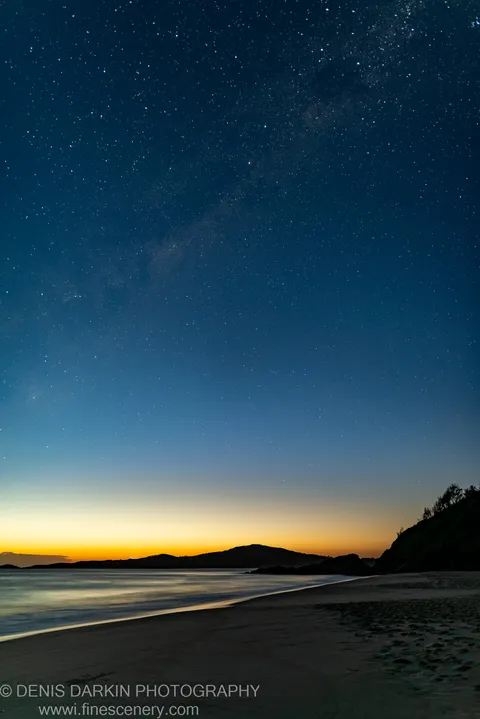 The Milky Way arches over the Nelson Bay coastline before dawn, with amber light on the horizon and long-exposure water along the shore.