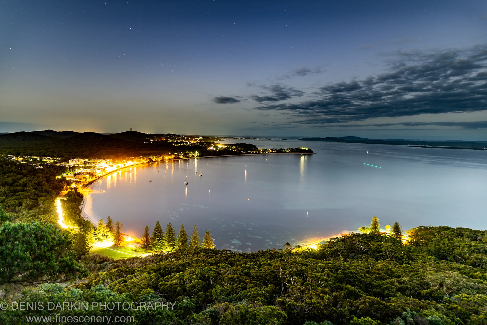 Coastal vegetation overlooks Nelson Bay at night, with amber town lights reflected across still water and a gradient blue sky above.