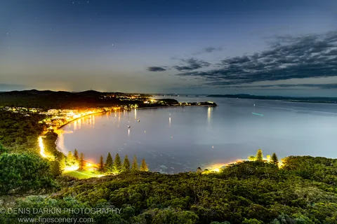 Coastal vegetation overlooks Nelson Bay at night, with amber town lights reflected across still water and a gradient blue sky above.