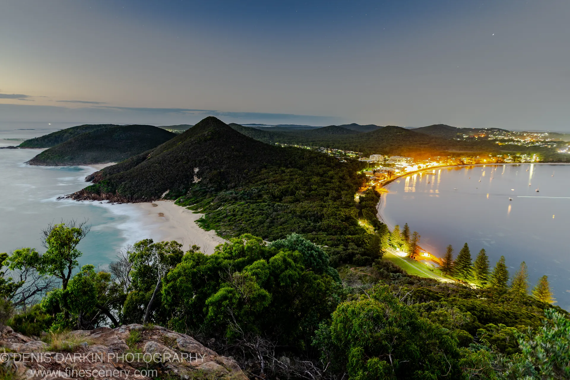 Long exposure at blue hour over Nelson Bay harbor, with smooth surf and calm reflective water below the forested headland.