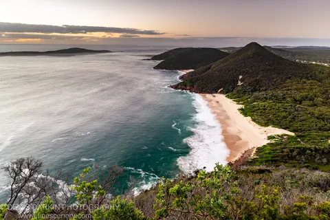 Long-exposure dawn view of a curved coastal bay at Nelson Bay, pale surf breaking against white sand below scrub-covered hills.