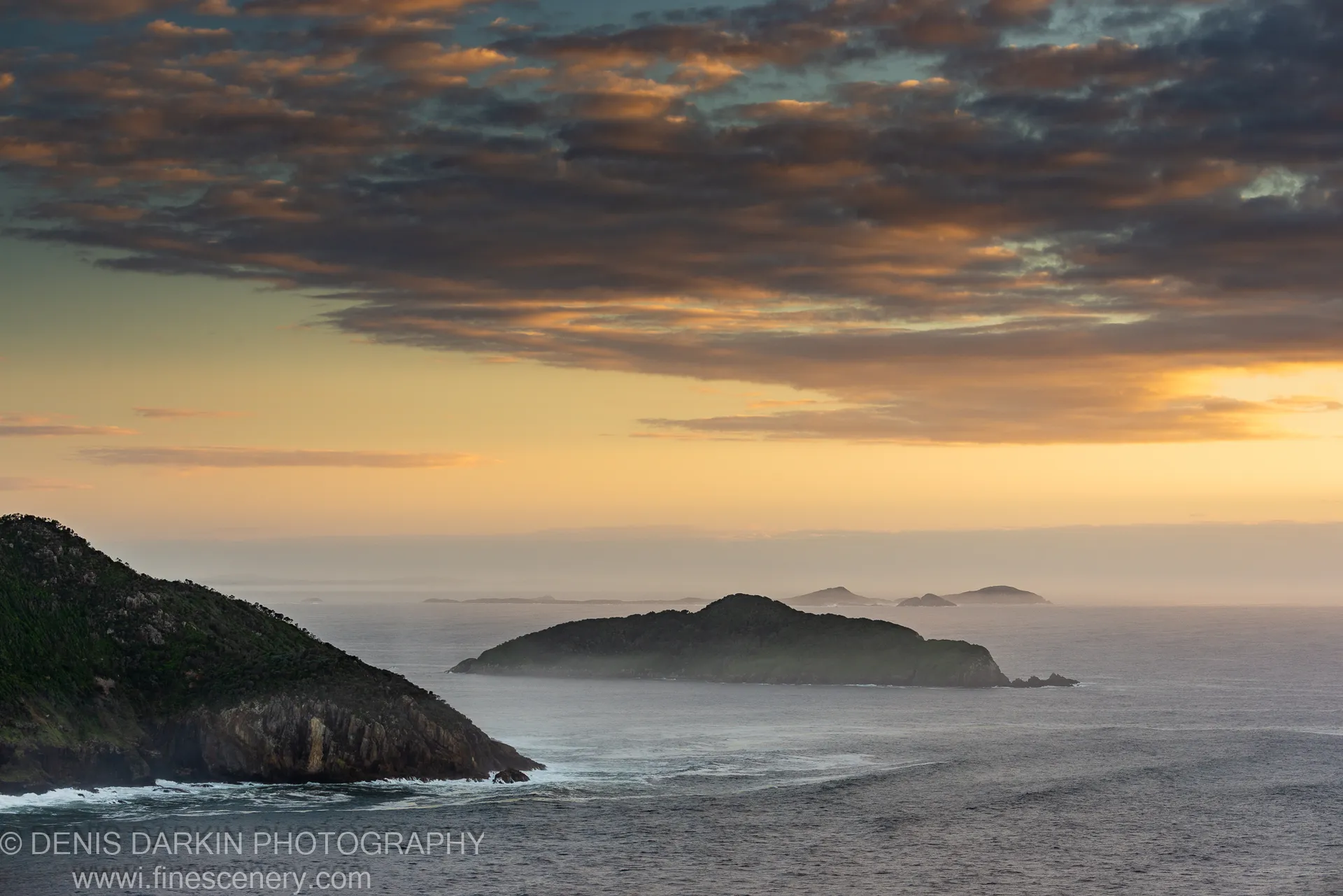 Coastal dawn view from Nelson Bay headland, with amber light on the horizon and mist-shrouded islands across grey water.