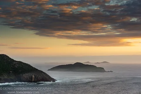 Coastal dawn view from Nelson Bay headland, with amber light on the horizon and mist-shrouded islands across grey water.