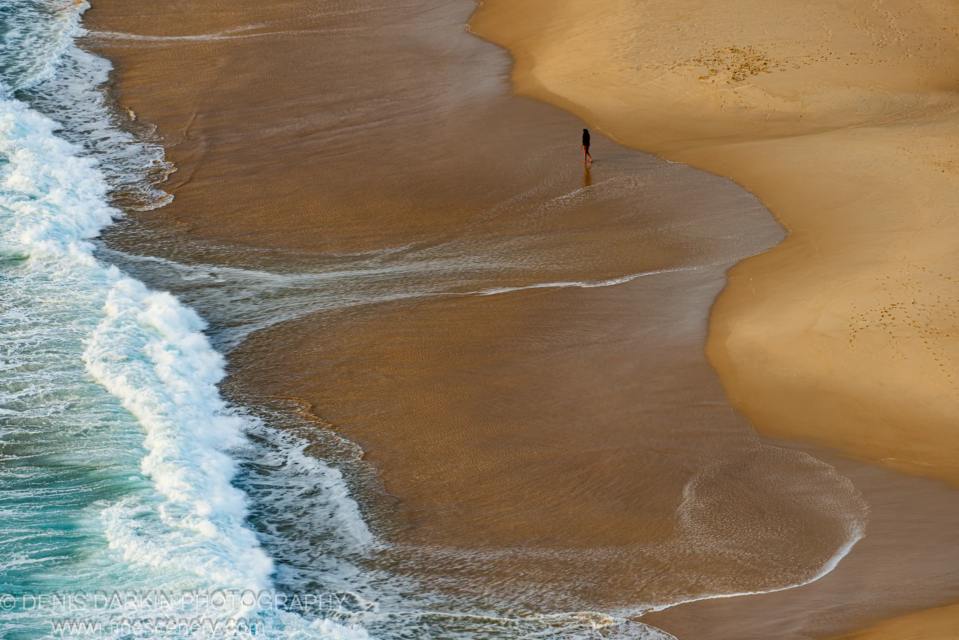 A lone figure walks the wide tidal flat at Nelson Bay, wet sand reflecting warm early light against incoming surf.