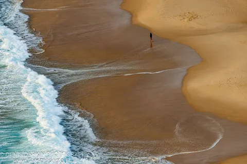 A lone figure walks the wide tidal flat at Nelson Bay, wet sand reflecting warm early light against incoming surf.