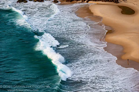 Telephoto view of a solitary figure walking the surf line at Nelson Bay, white foam tracing the curve of the beach at dawn.