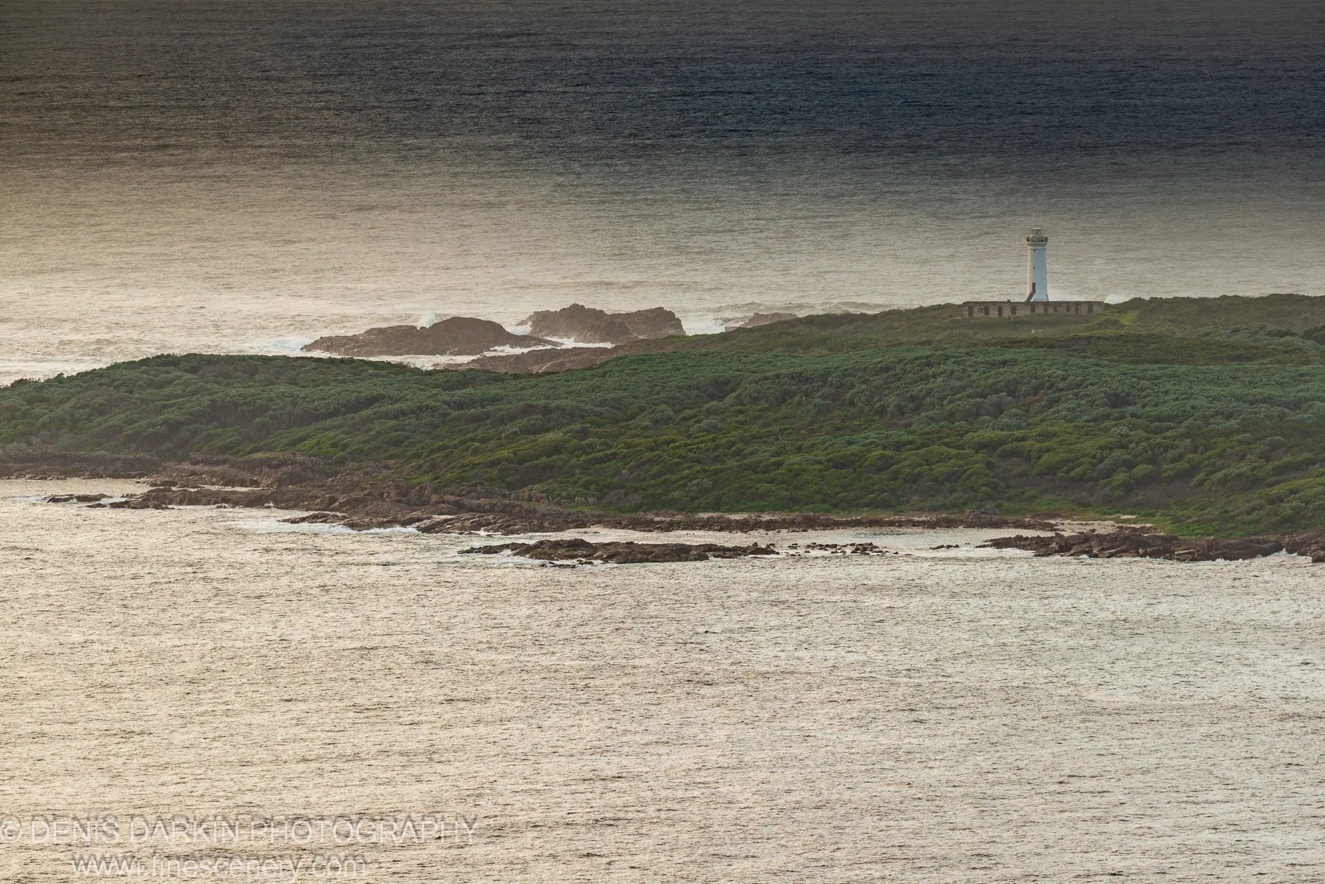 A white lighthouse tops a scrub-covered headland above dark rocks and sea foam, Nelson Bay coast.