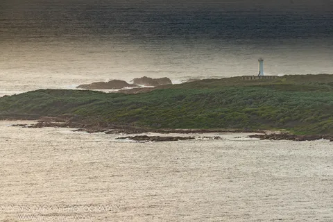 A white lighthouse tops a scrub-covered headland above dark rocks and sea foam, Nelson Bay coast.