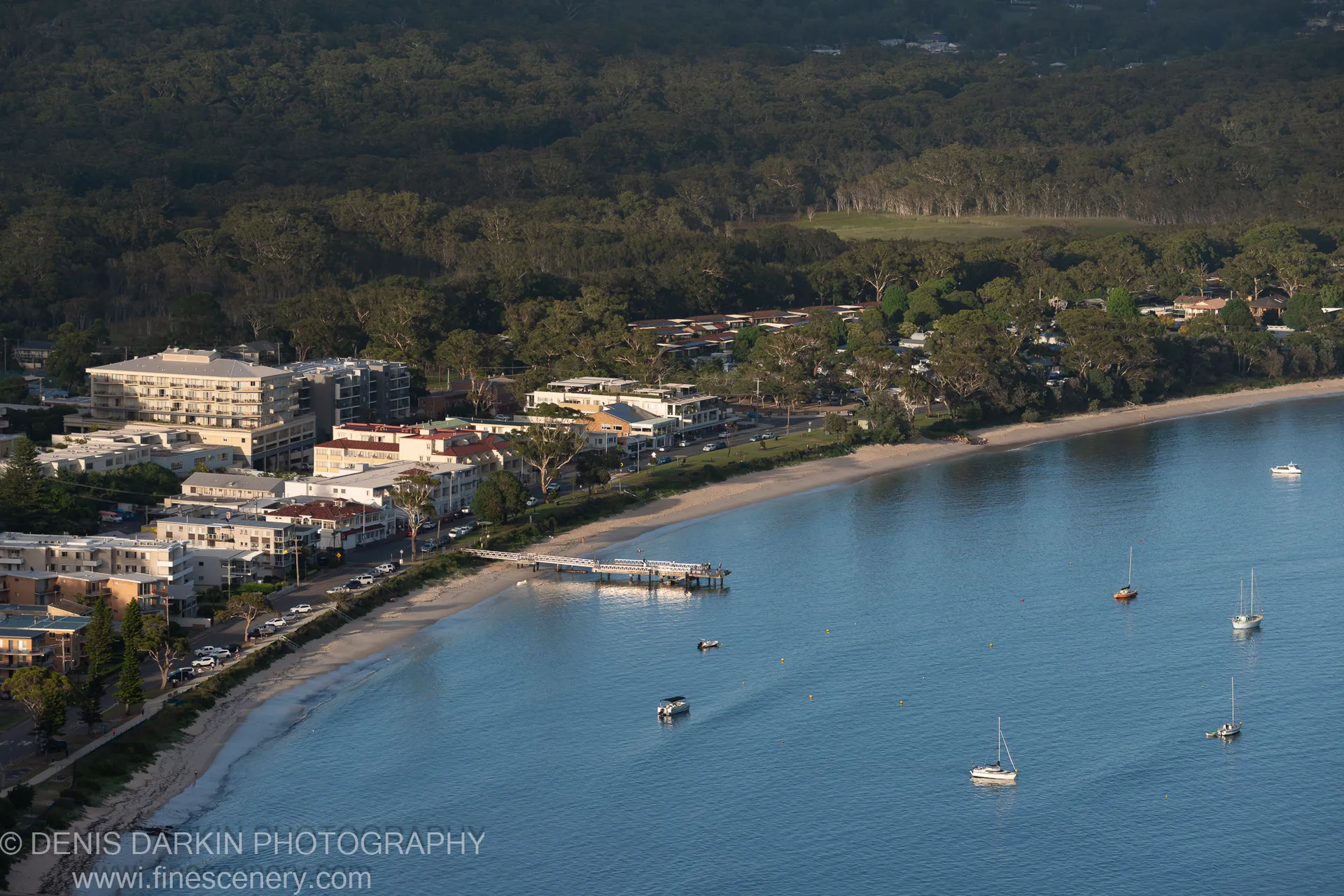 Waterfront buildings and sailboats along the sandy shore of Nelson Bay on a summer morning, with a wooden pier extending into the calm blue bay.