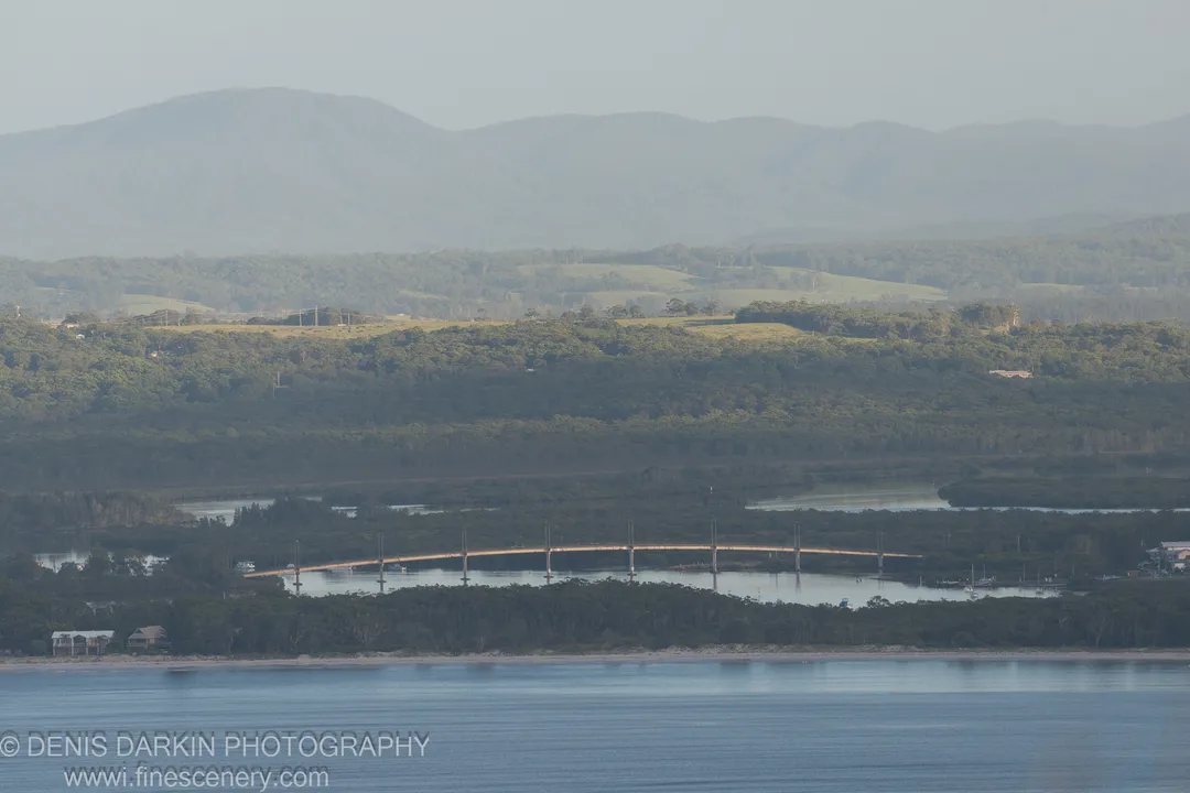 A long concrete bridge spans a waterway bordered by dark mangrove vegetation at Nelson Bay, with rolling hills and hazy blue mountains in the distance.