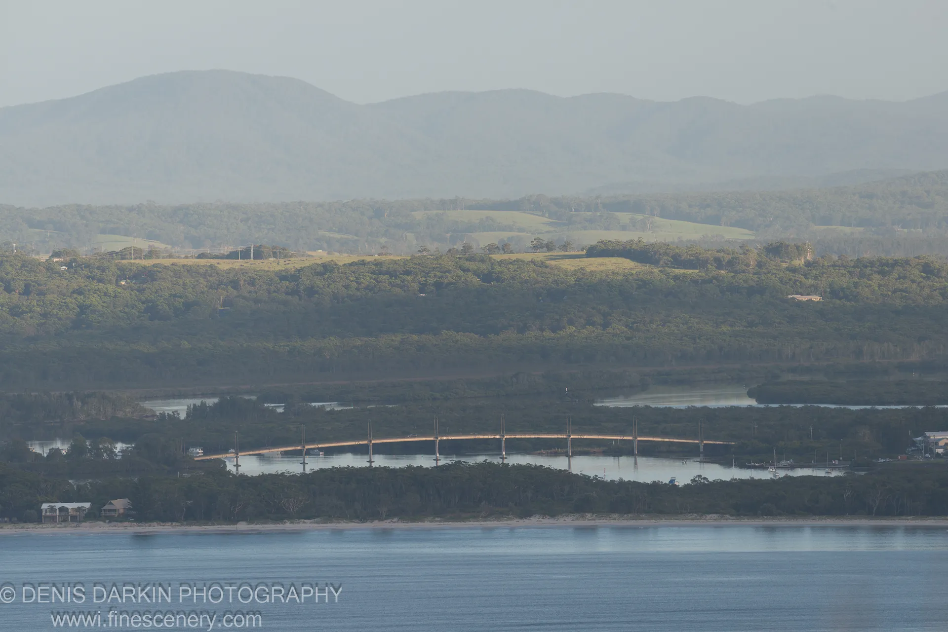 A long concrete bridge spans a waterway bordered by dark mangrove vegetation at Nelson Bay, with rolling hills and hazy blue mountains in the distance.