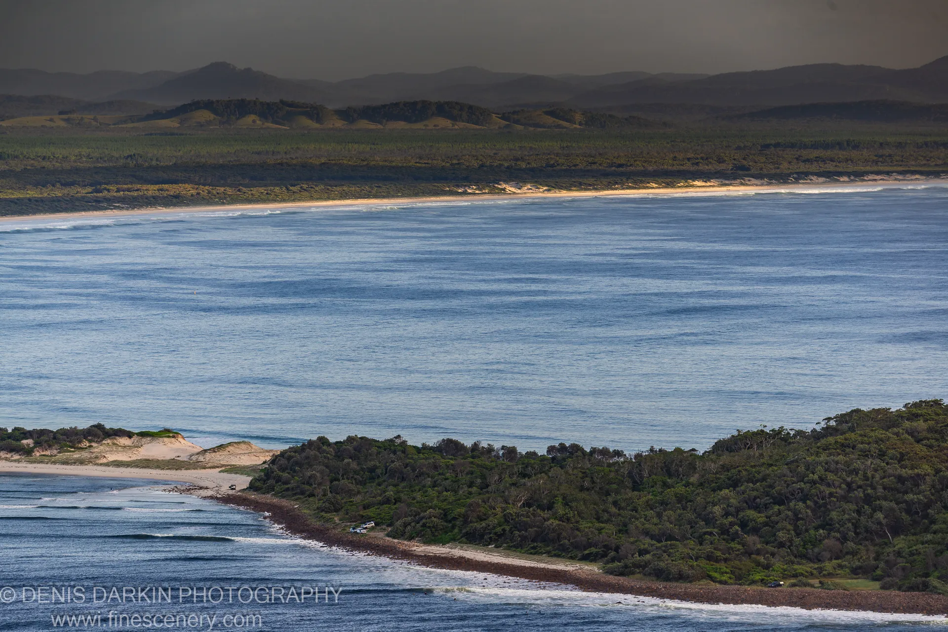 Dark rocky shoreline leads to deep blue water and sunlit hills across Nelson Bay, shadowed forest on the near bank.