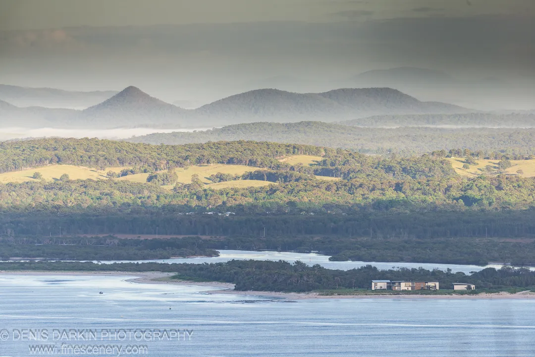 Telephoto compression layers rolling green hills above a calm estuary at Nelson Bay, hazy mountains behind a sandy spit and low buildings.