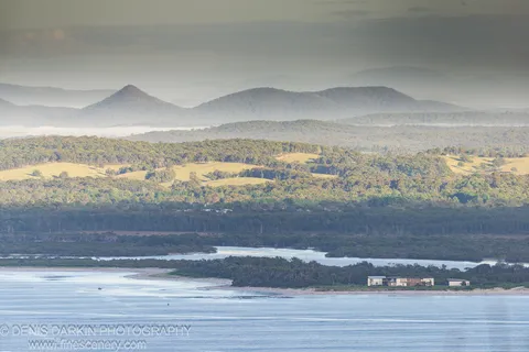 Telephoto compression layers rolling green hills above a calm estuary at Nelson Bay, hazy mountains behind a sandy spit and low buildings.