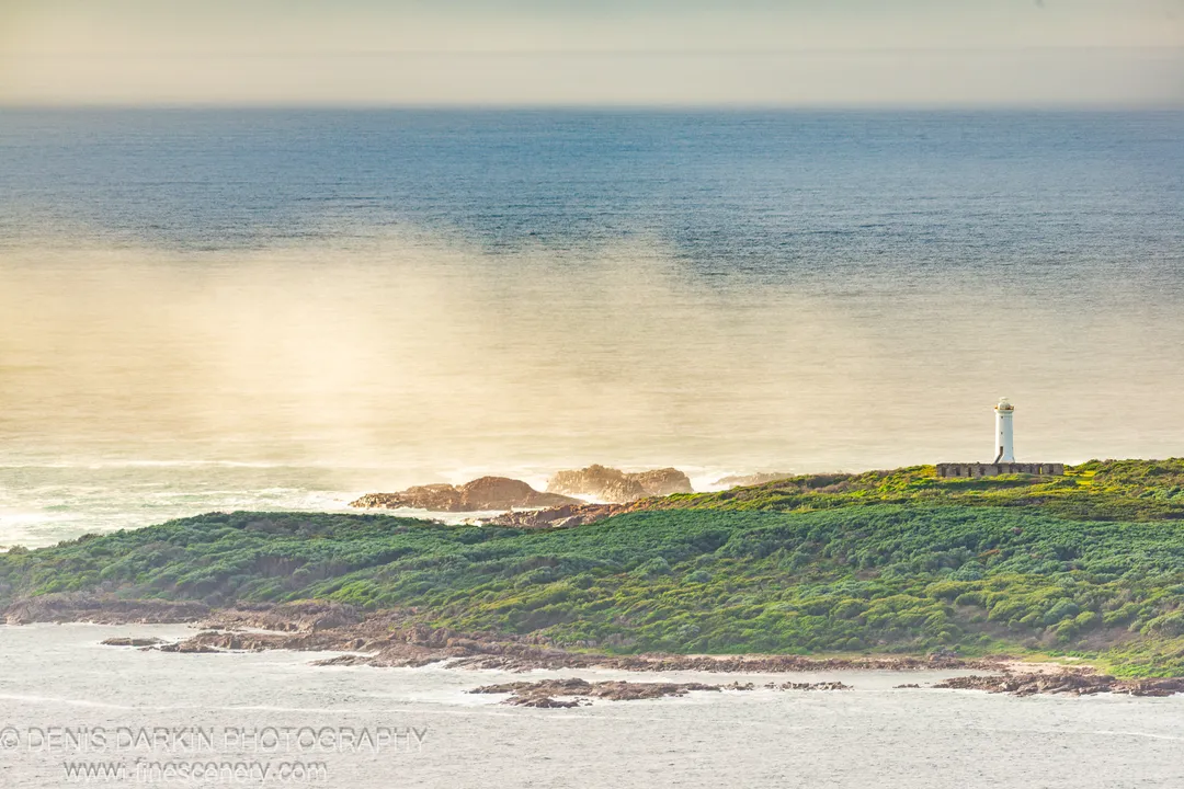 White lighthouse on a rocky green headland at Nelson Bay, New South Wales, with sunlight reflecting off the ocean surface below.