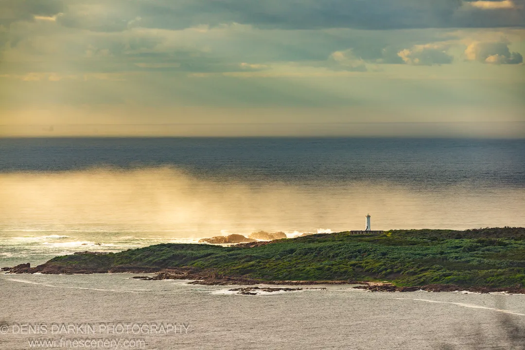 The lighthouse at Nelson Bay rises above a scrub-covered coastal headland with golden light breaking through heavy cloud overhead.