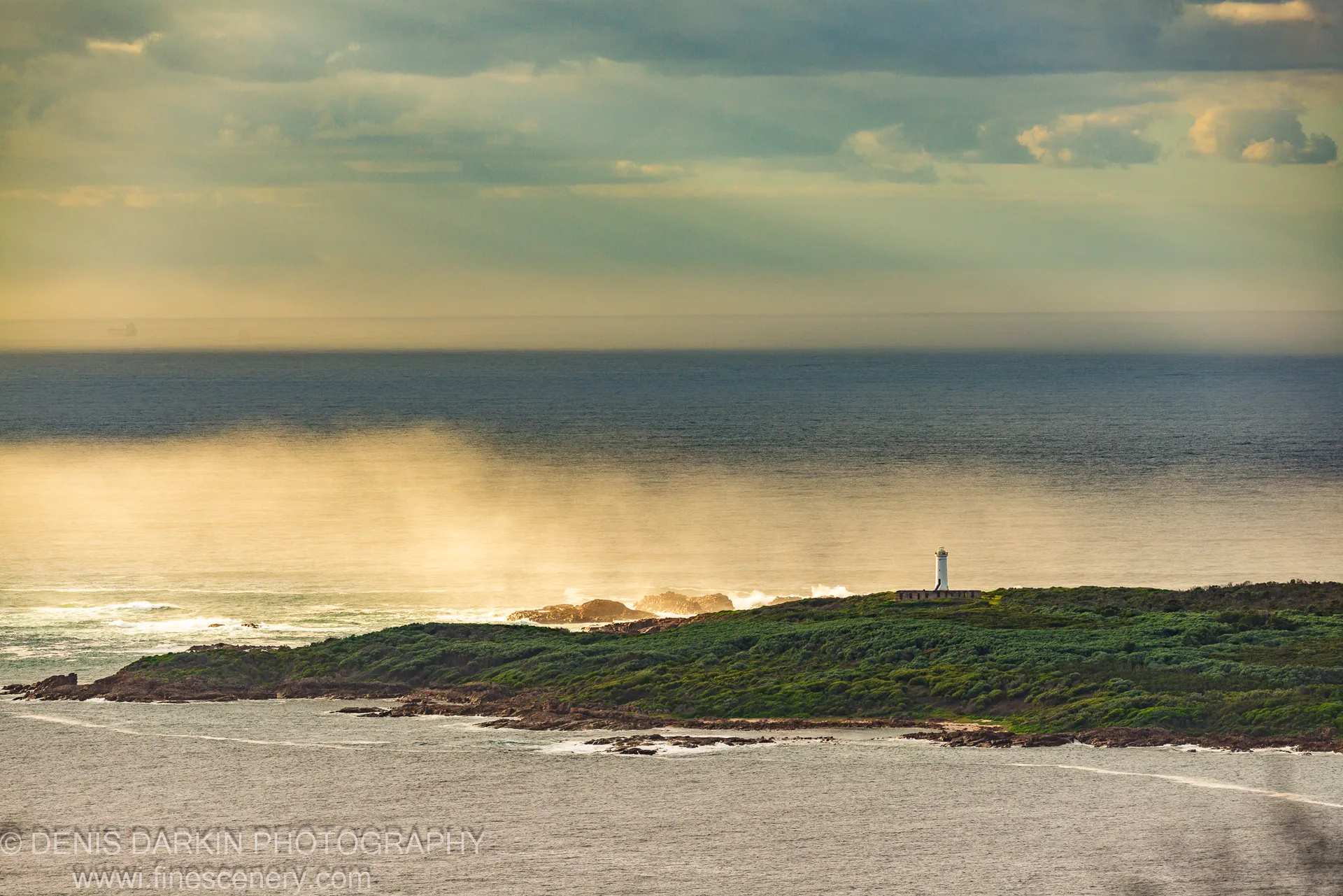 The lighthouse at Nelson Bay rises above a scrub-covered coastal headland with golden light breaking through heavy cloud overhead.