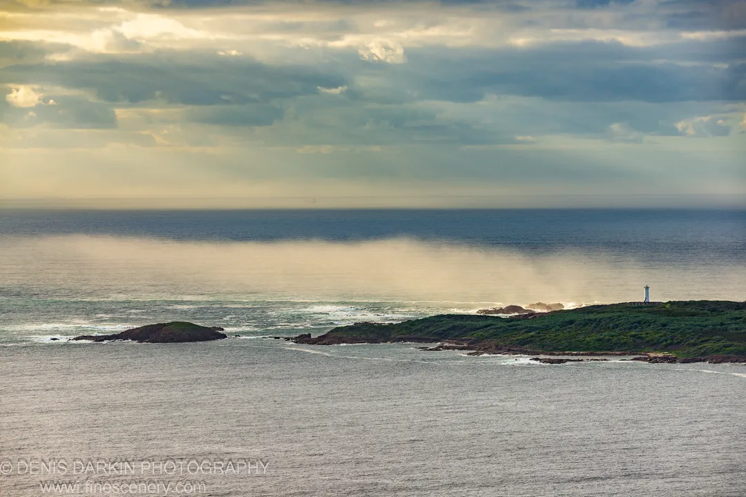 Rays of sunlight pass through heavy cloud above the rocky headland at Nelson Bay, with the white lighthouse visible against the grey sky.