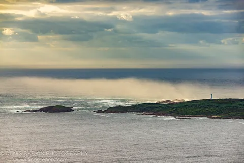 Rays of sunlight pass through heavy cloud above the rocky headland at Nelson Bay, with the white lighthouse visible against the grey sky.