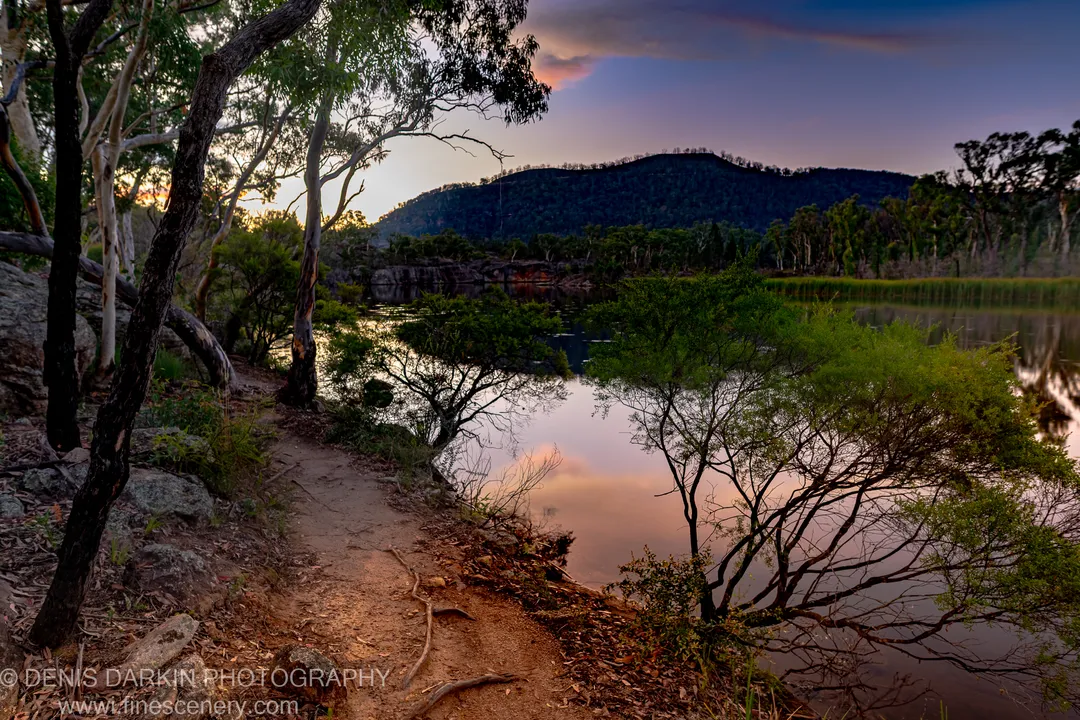 A winding dirt track curves along the riverbank at dusk, with silhouetted hills against a twilight sky reflected in calm water, Dunns Swamp.
