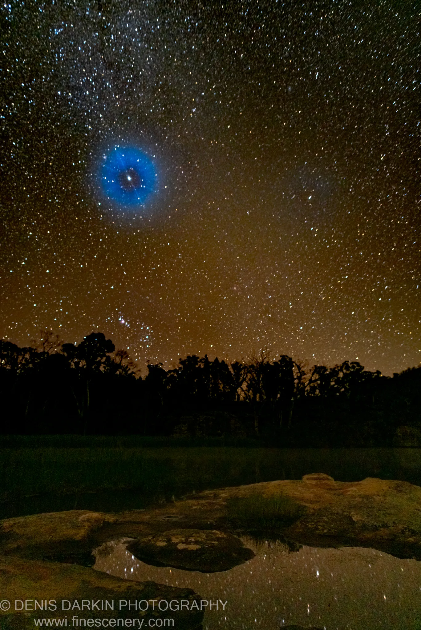 A blue planetary nebula hangs low in the night sky above silhouetted trees at Dunns Swamp, with rocky shallows and still pools below reflecting the stars.