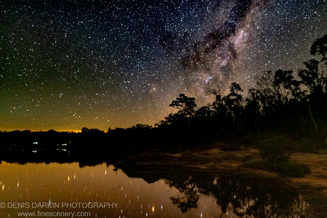 The Milky Way spans the sky above silhouetted trees at Dunns Swamp, an orange glow along the horizon mirrored in still water below.