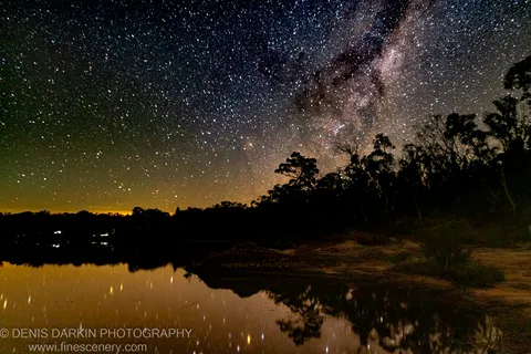 The Milky Way spans the sky above silhouetted trees at Dunns Swamp, an orange glow along the horizon mirrored in still water below.