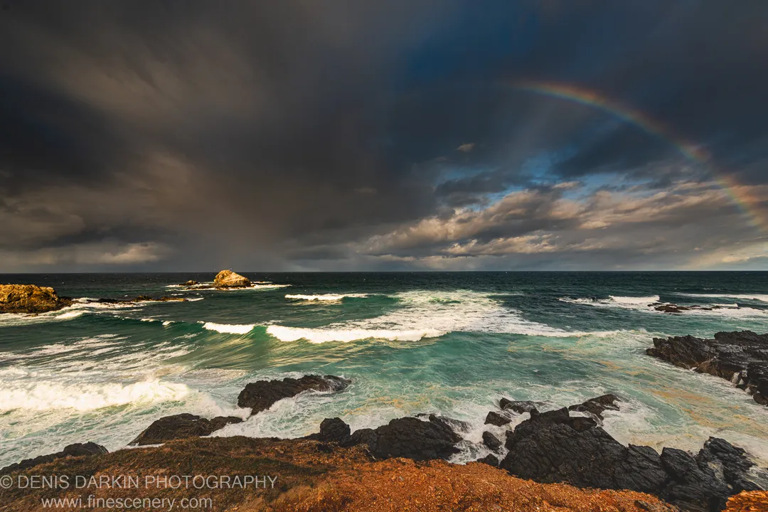 A rainbow arcs above storm clouds and teal surf breaking over dark rock at the Byron Bay coast, New South Wales.