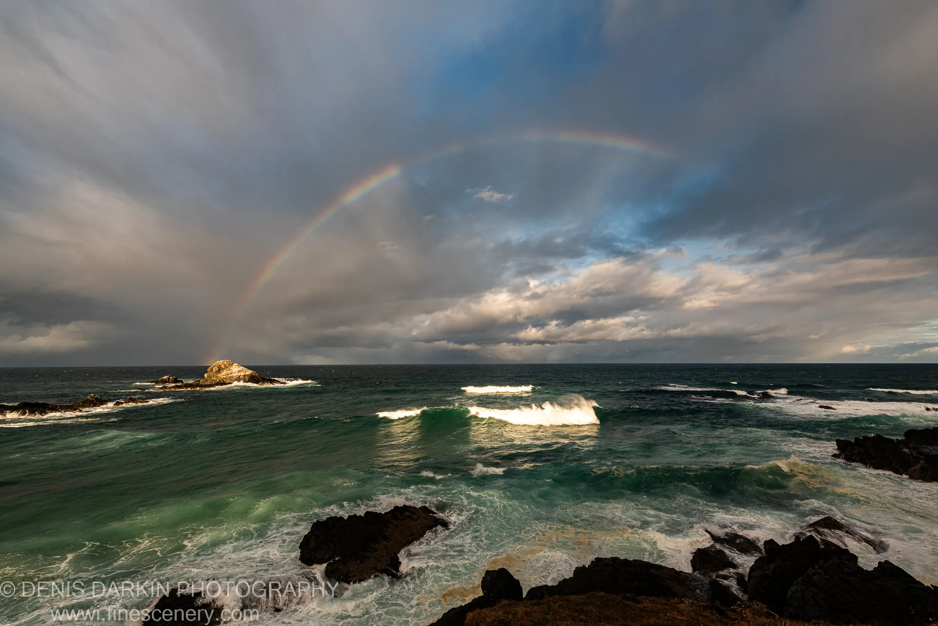 A full rainbow arcs from dark storm clouds to the horizon above surf crashing over jagged rocks at Byron Bay, New South Wales.