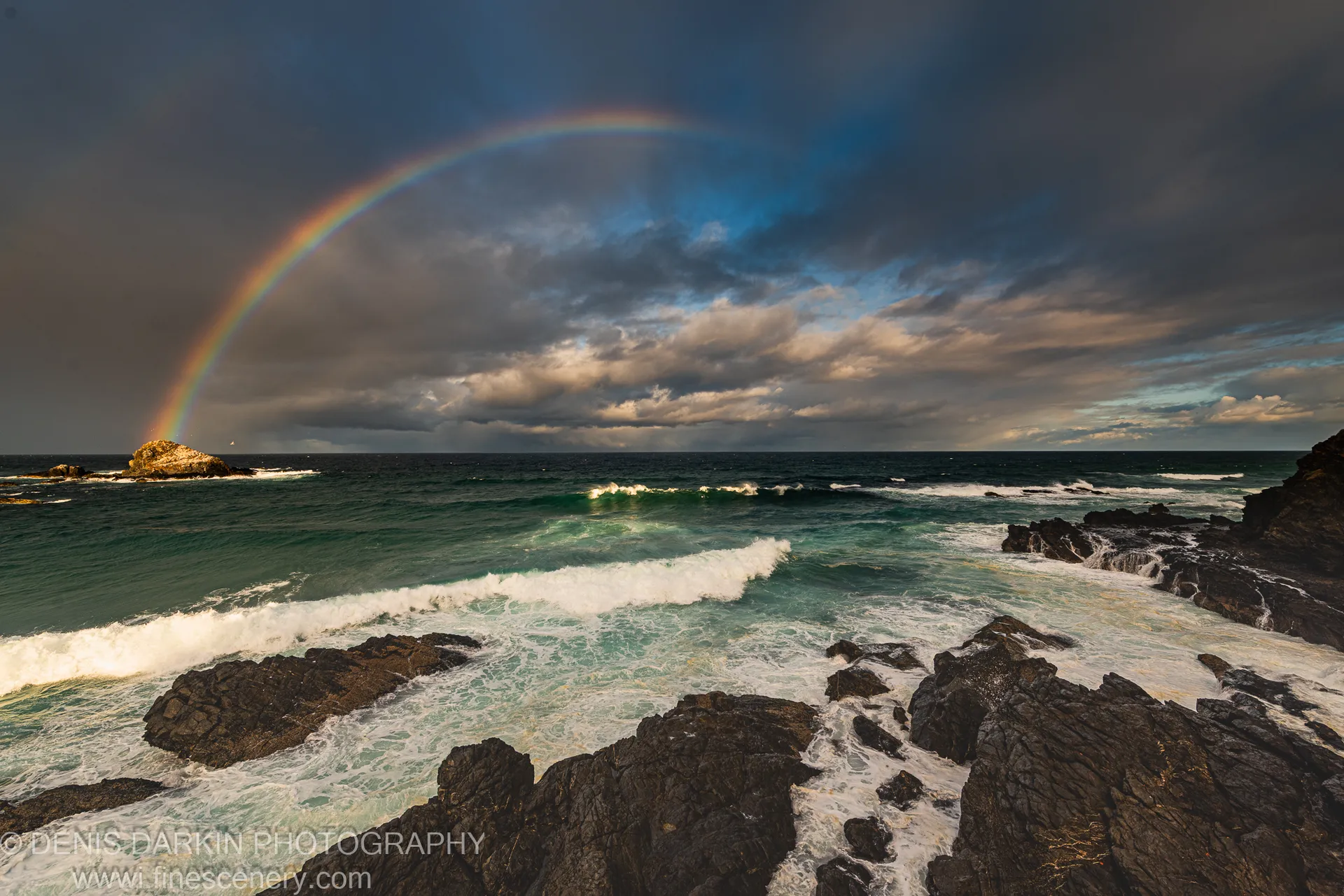 A full-arc rainbow stretches over dark basalt rock formations and a sea stack along the Tallow Beach coast in winter.