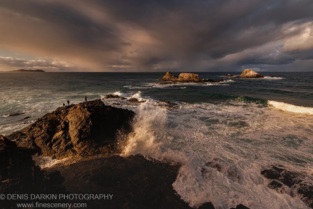 Warm golden-hour light on sea stacks and dark wet rocks as waves crash against a rugged NSW headland at Byron Bay.