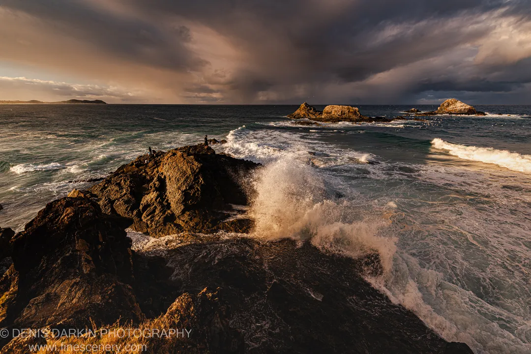 A wave explodes against a dark headland at Byron Bay, sending a tall plume of spray skyward while figures stand on the rocky outcrop.