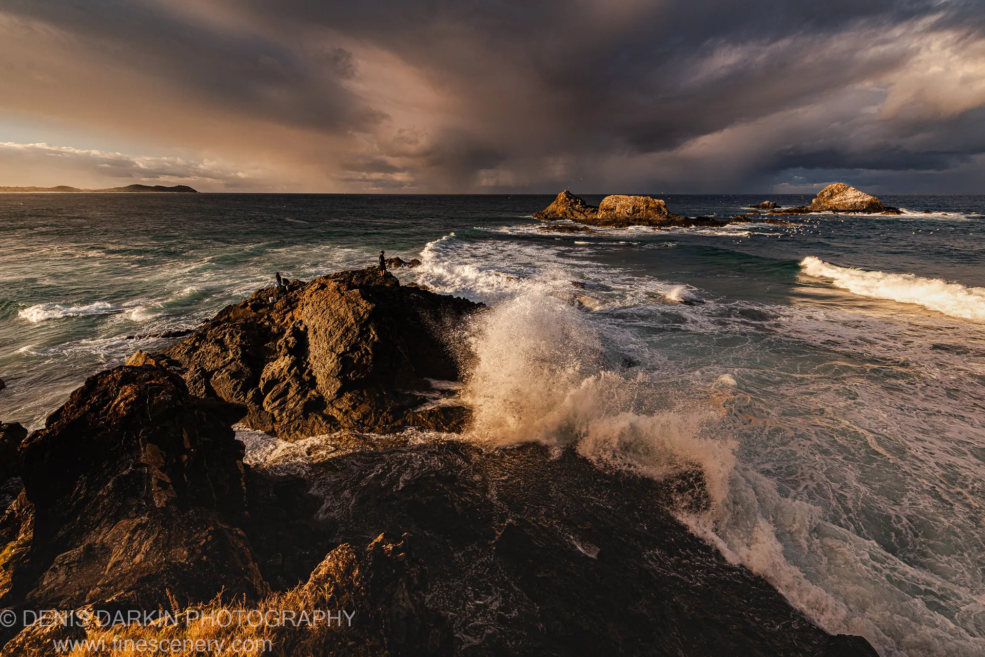 A wave explodes against a dark headland at Byron Bay, sending a tall plume of spray skyward while figures stand on the rocky outcrop.